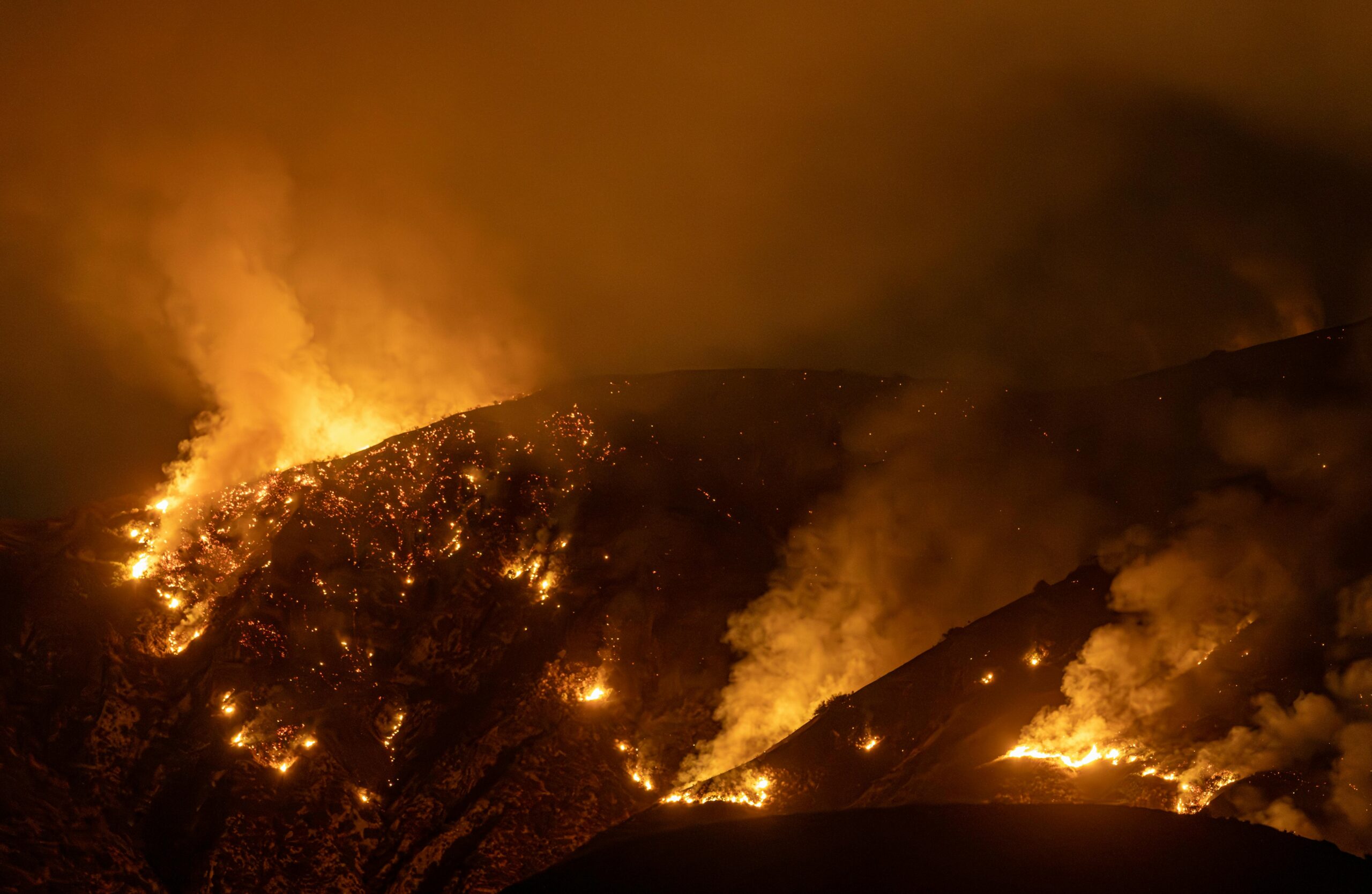 Intense wildfire burning on a California hillside at night, producing heavy smoke and ash.